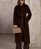 A woman wears this longline coat featuring oversized pockets perfect for sinking hands into, with a generous collar and lapels that create a bold statement. She styles it with the matching thick faux fur belt, cinching the waist for a flattering silhouett