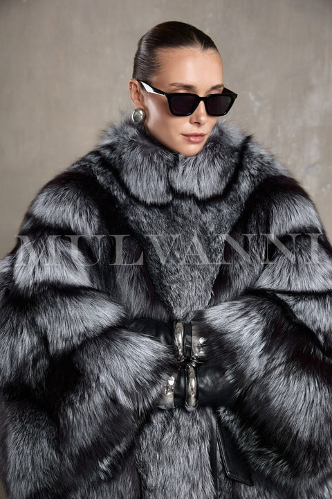 A woman wears an elegant full-length silver fox fur coat with a voluminous silhouette and leather belt that cinches the waist.