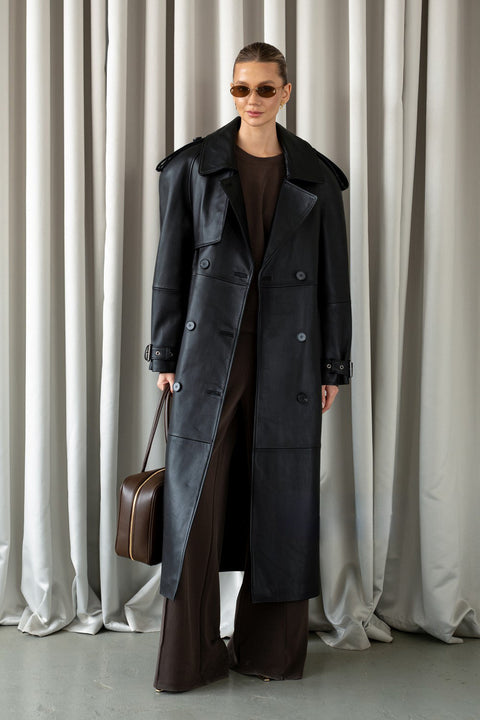 Elegantly dressed, a woman models a black leather trench coat with a classic double-breasted design, sophisticated notched lapels, and belted cuffs made from premium soft leather.