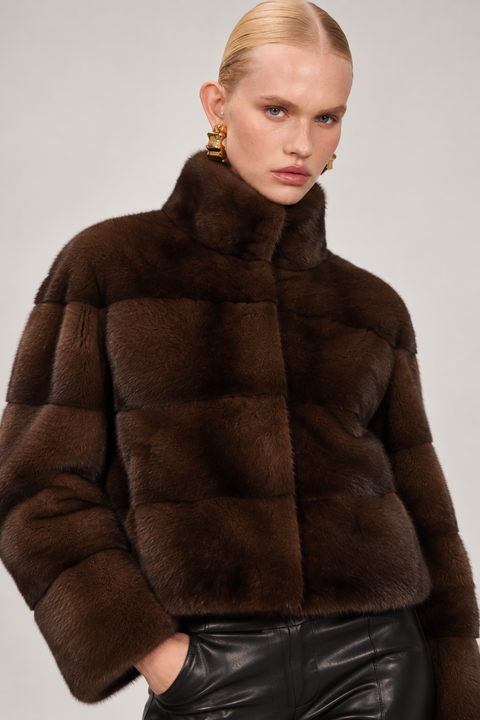 A model wearing a cropped, dark brown fur jacket with a high collar and black leather pants, photographed against a neutral light gray studio background.